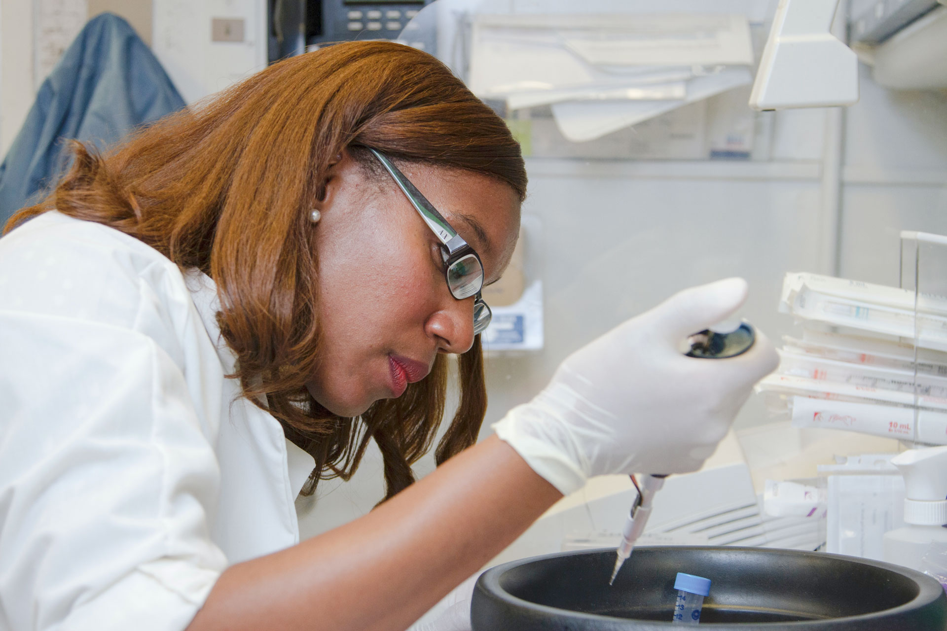 Black woman wearing glasses, leans over a vial whilst holding a pipette