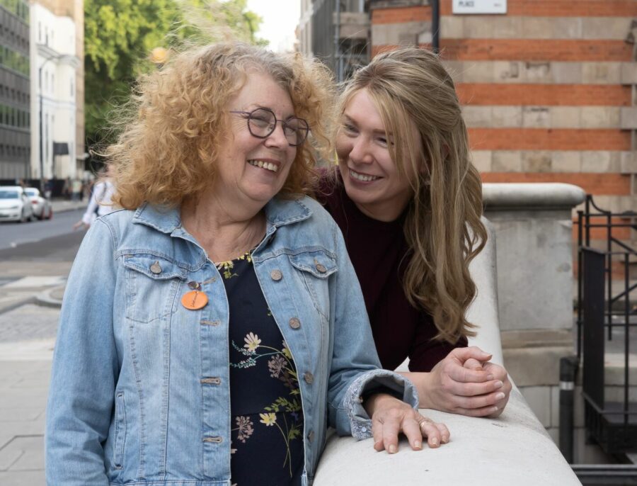 A Mum where leukaemia UK pin badge and adult daughter leaning on a wall and smiling together on a city street