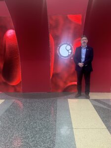 Dr Simon Ridley stood in a suit at the ASH conference next to a red wall with the ASH logo