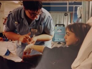 Jesse in a hospital bed with a nurse injecting her bandaged hand pre transplant