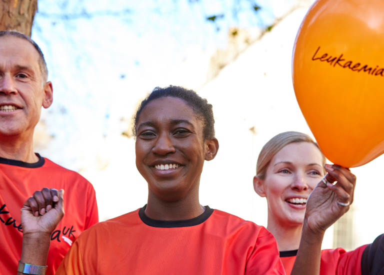3 people outside where Leukaemia UK T-Shirts. The person in the front smiling holding both hands up and in their left hand is holding an orange Leukaemia UK balloon