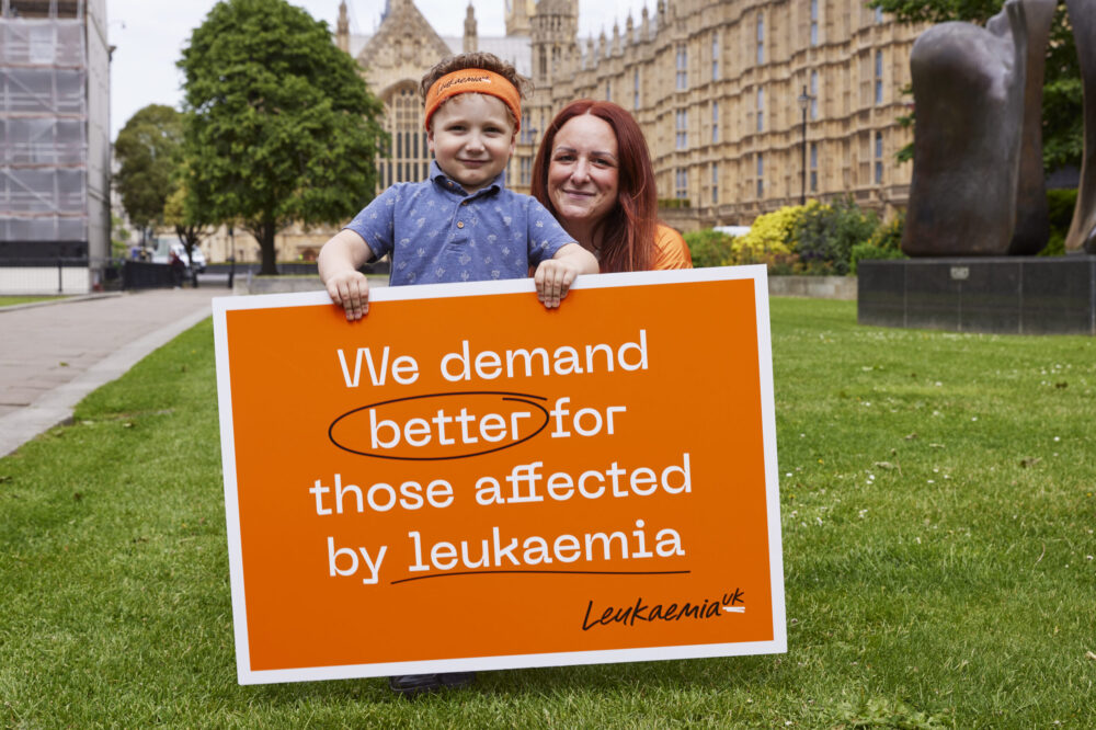 Mother and young son outside houses of Parliament with a Leukaemia UK campaigning board that says 'we demand better for those affected by leukaemia'