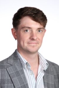 Headshot of Dr Shaun Patterson, John Goldman Fellowship awardee 2026, in a grey check suit jacket and striped white shirt